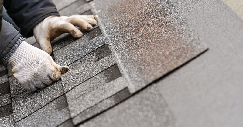 Close up and real photo of skilled workman in special protective work wear installing asphalt or bitumen tile on top of the roof under construction house